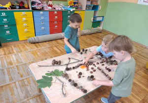 Trzech chłopców stoją przy stoliku i tworzą pejzaż jesienny z materiałów przyrodniczych.