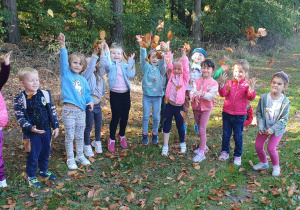 Dzieci z grupy Słoneczka stoją na ścieżce leśnej podrzucając jesienne liście.