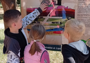 Troje przedszkolaków stoi przed tablicą edukacyjną pt.:” Kopalnia węgla kamiennego”. Dzieci oglądają ilustrację, chłopiec po lewej stronie wskazuje na tablicę palcem tłumacząc pozostałym proces wydobycia węgla.