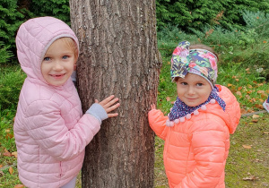 Dwie dziewczynki z grupy Liski pozują do zdjęcia przytulając się do drzewa.