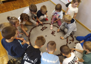 Dzieci układają na dywanie uśmiechniętą buźkę z kasztanów.