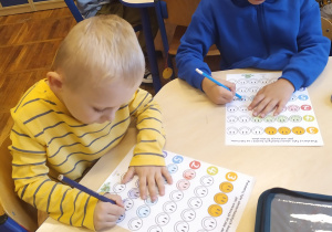Dwóch chłopców siedzi przy stoliku i koloruje kartę pracy – tyle uśmiechniętych buziek ile wskazuje cyfra obok.