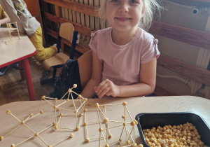 Dziewczynka siedzi przy stoliku i prezentuje swoją konstrukcję.