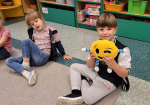 Chłopiec siedzi z dziewczynką na dywanie i trzyma maskotkę (uśmiechniętą buźkę).