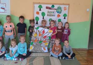 Dzieci z grupy „Misie” pozują do zdjęcia na tle tablicy dekoracyjnej w rękach trzymają pracę plastyczną „Drzewko”, które wykonały z szarego papieru na dużym białym brystolu i przykleiły liście ze swoimi imionami.