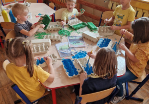 Dzieci siedzą przy stolikach i malują farbami swoje wytłaczanki.