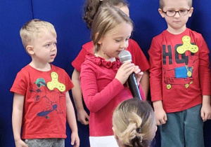 Dziewczynka w czerwonej bluzce mówi wierszyk przez mikrofon.