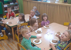 Dzieci siedzą przy stolikach i przed sobą mają talerzyki na których leży chleb.