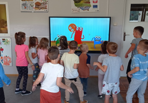 Wykorzystanie monitora podczas zajęć ruchowych- grupa dzieci stoi przodem do tablicy i wykonuje ruchy gimnastyczne prezentowane w piosence.