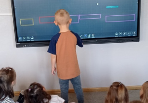 Wykorzystanie monitora podczas zajęć z prostokątami- Chłopiec w pomarańczowej bluzce rysuje na ekranie swój prostokąt.