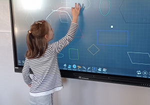 Wykorzystanie monitora podczas zajęć z prostokątami- dziewczynka w bluzce w paski łączy dwie takie same figury geometryczne rysując linie palcem po ekranie.